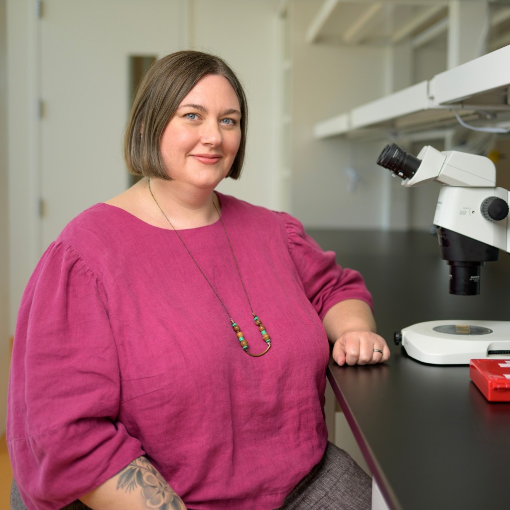 Photo of a woman in a pink blouse sitting near a microscope in a lab setting.