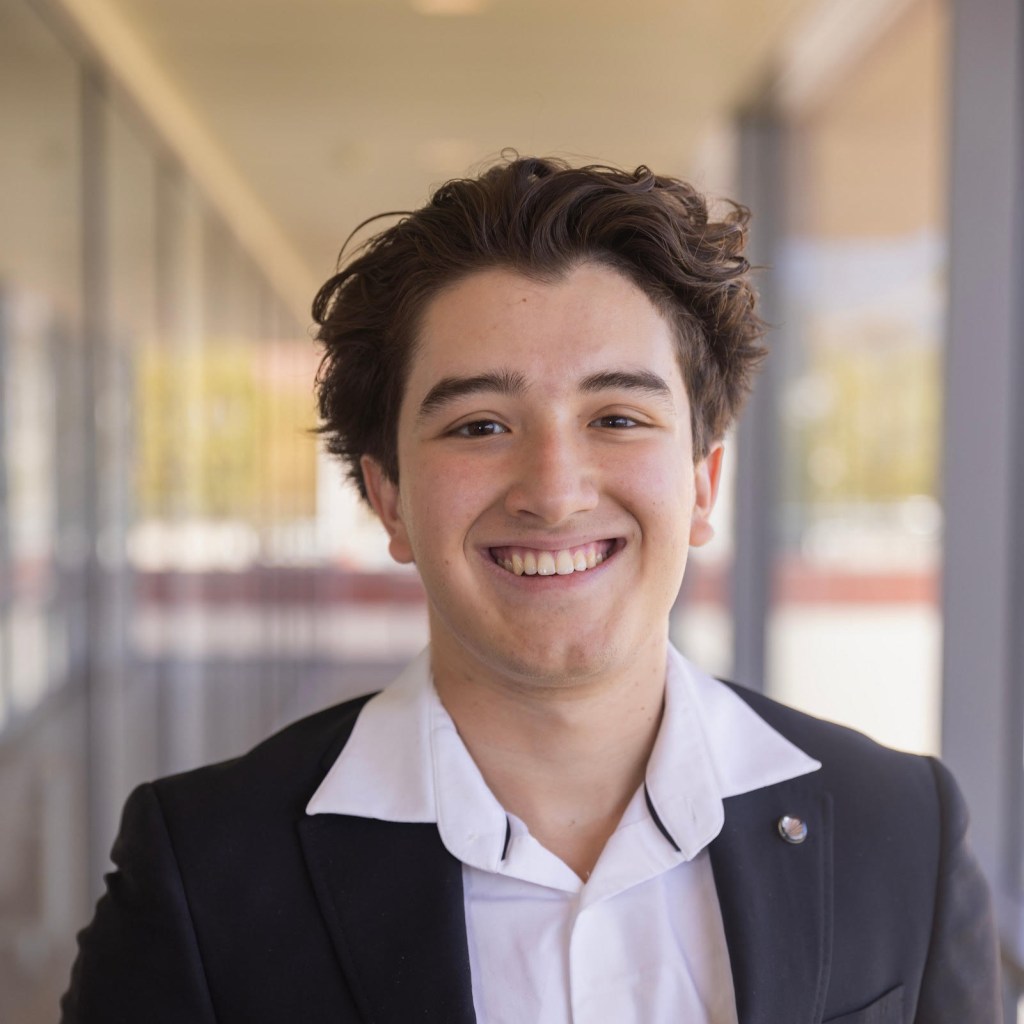 Young man smiling while wearing a black suit jacket and white shirt, standing in a corridor with natural light.