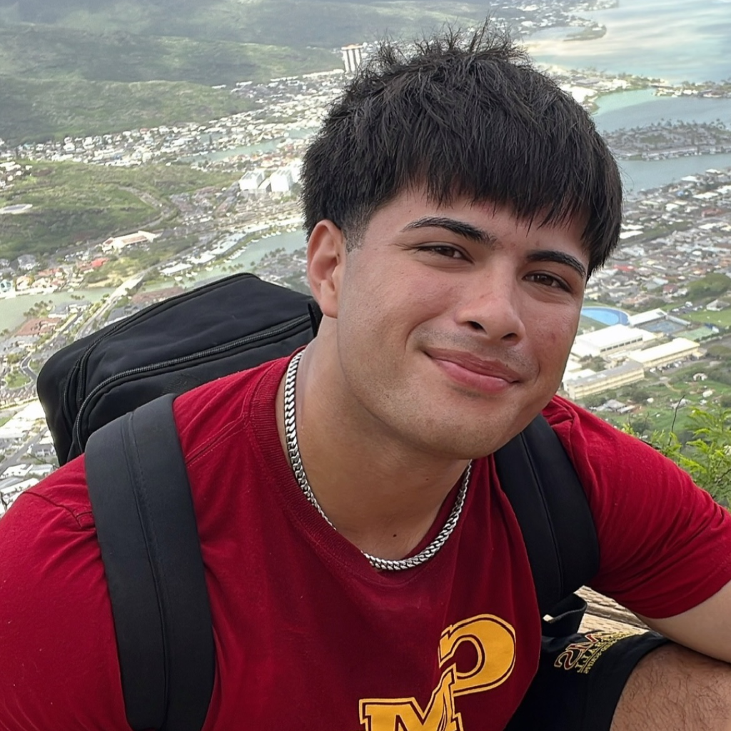 A smiling young man with short dark hair, wearing a red shirt and a silver necklace, poses outdoors with a scenic landscape visible behind him.