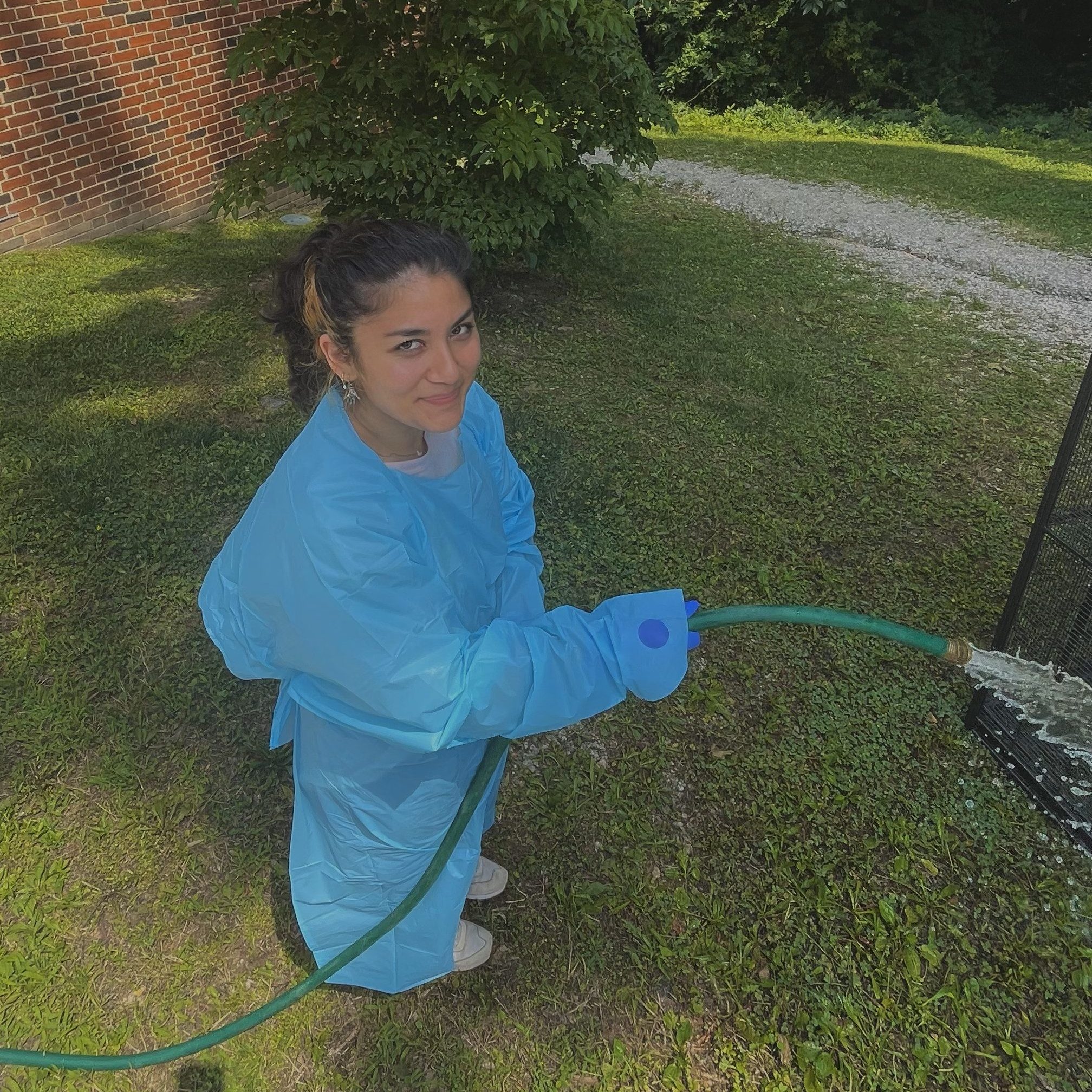 A laboratory technician wearing a blue lab coat and gloves, using a garden hose to wash equipment in a grassy outdoor area.