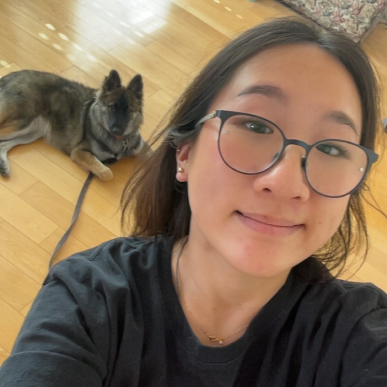 A young woman with glasses takes a selfie while sitting on a wooden floor, with a dog lying in the background.
