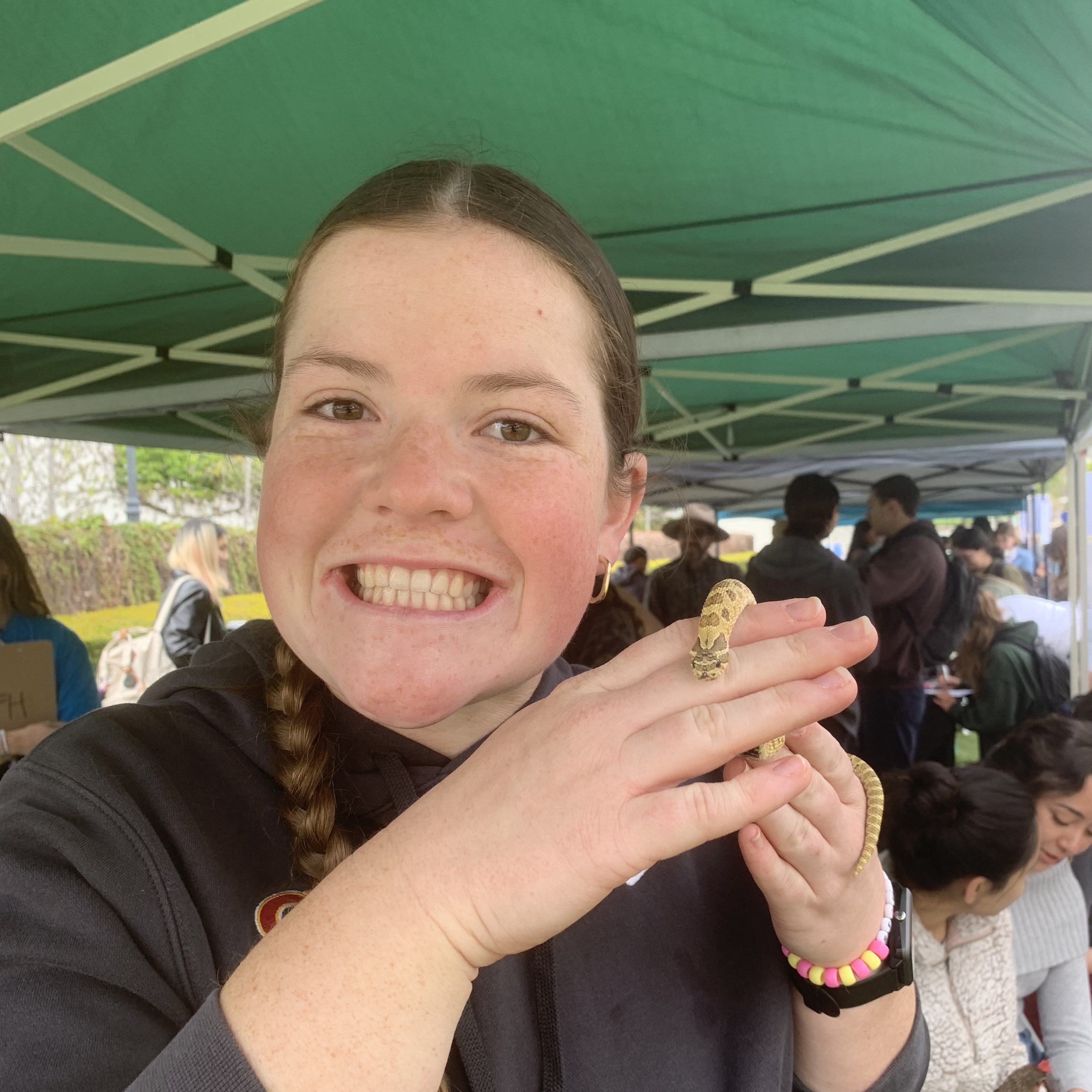 A smiling woman holding a small snake in her hands, standing under a green tent with people in the background.